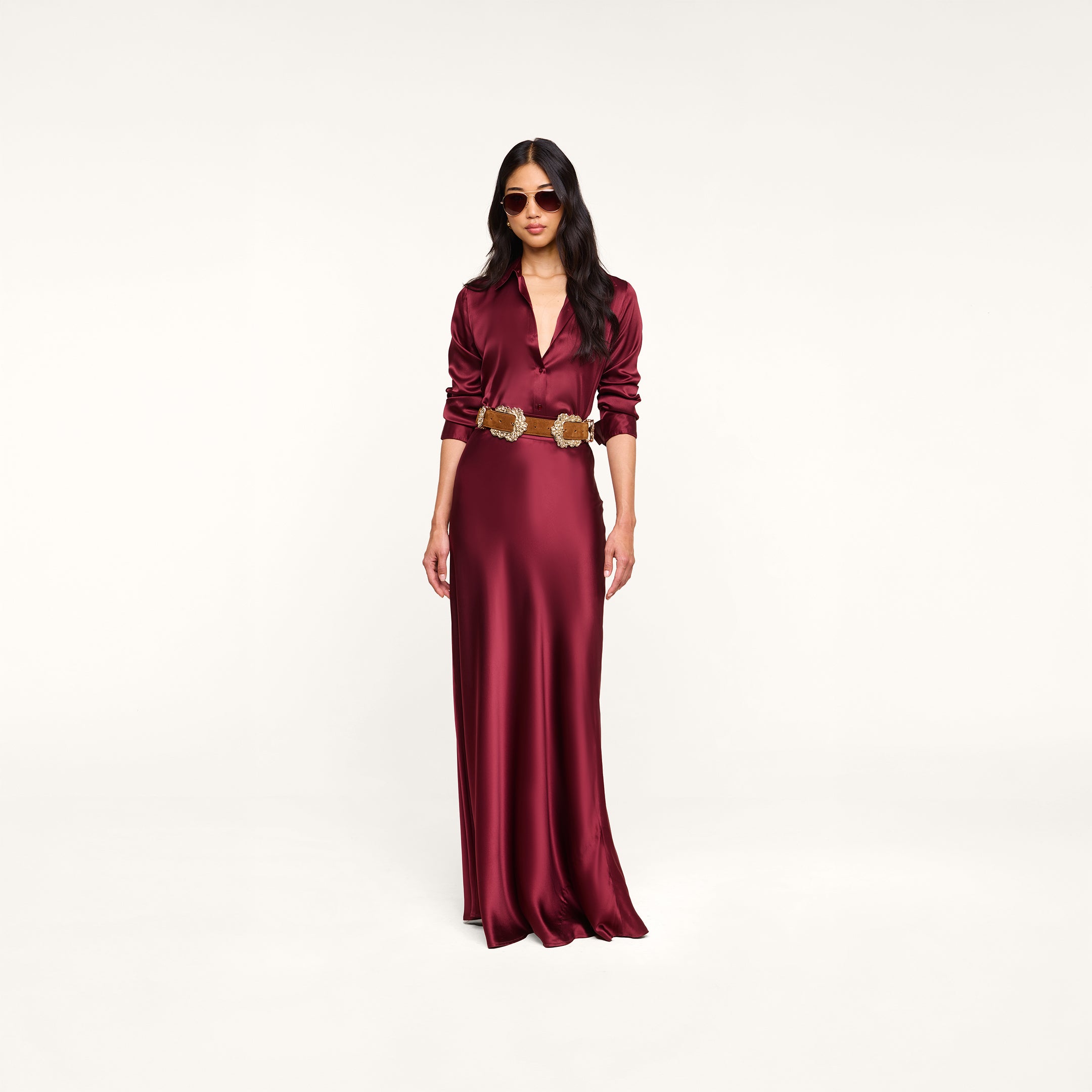 Wearing sunglasses, the woman stands against a plain white background in the Tyler Silk Blouse and a long, merlot red silk charmeuse skirt with a gold belt.
