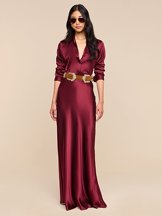 A woman wearing the Tyler Silk Blouse in merlot red, styled with a collared neckline, long sleeves, sunglasses, and a wide brown belt with decorative buckles, stands against a plain beige background.
