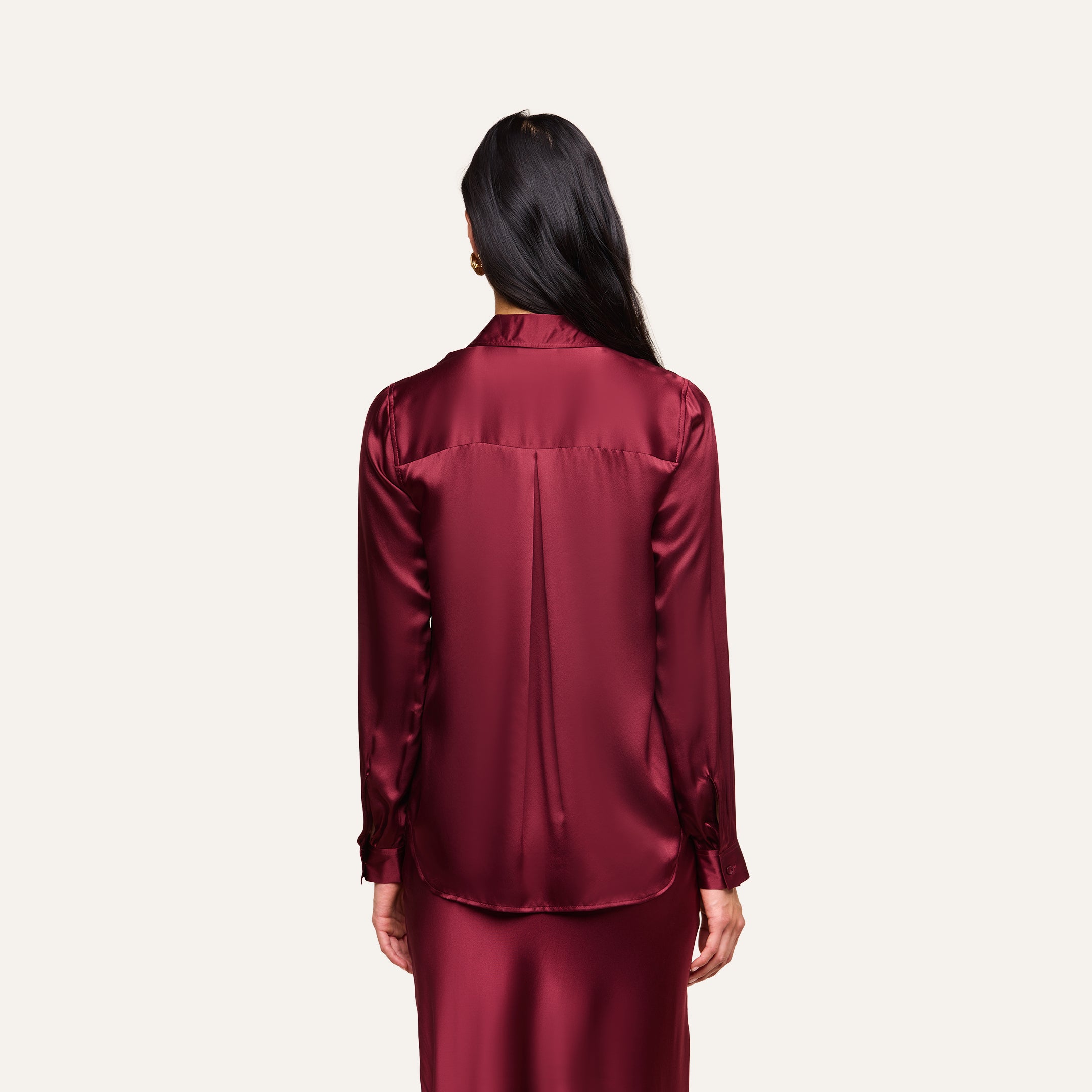 A woman with long dark hair, seen from behind, wears the Tyler Silk Blouse in merlot red along with a matching skirt against a plain light background.