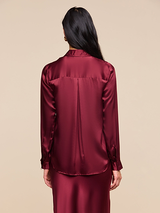 A person with long dark hair is shown from behind, wearing the shiny, long-sleeve Tyler Silk Blouse in merlot red along with a matching skirt, set against a plain beige background.