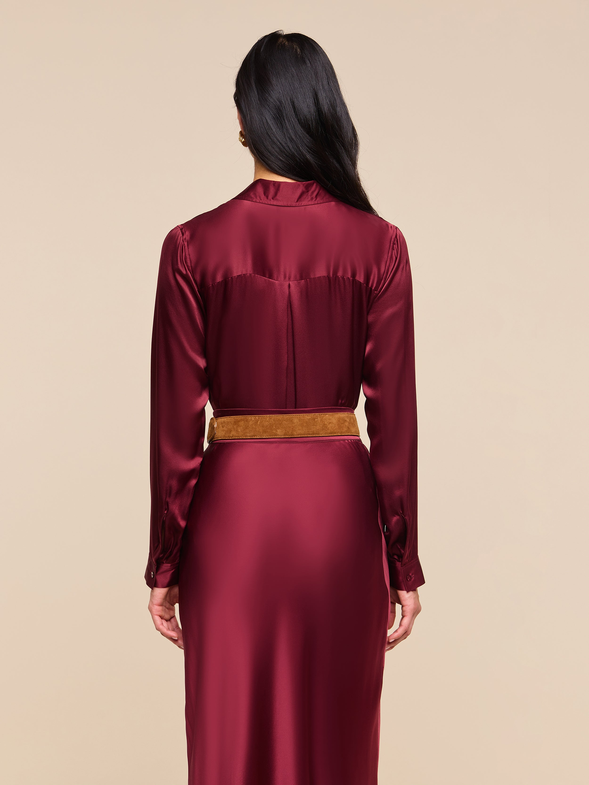 Woman with long dark hair stands facing away, wearing the Tyler Silk Blouse in merlot red with long sleeves and a tan belt, against an off-white background.