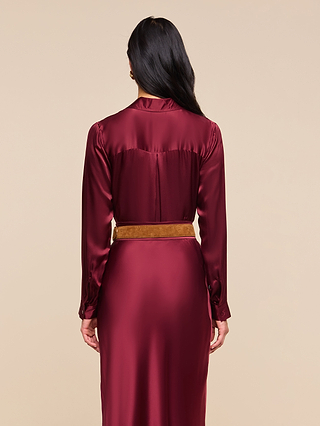 A woman with long dark hair wears the Tyler Silk Blouse in shiny merlot red, styled with a tan belt, standing with her back to the camera against a plain beige background.