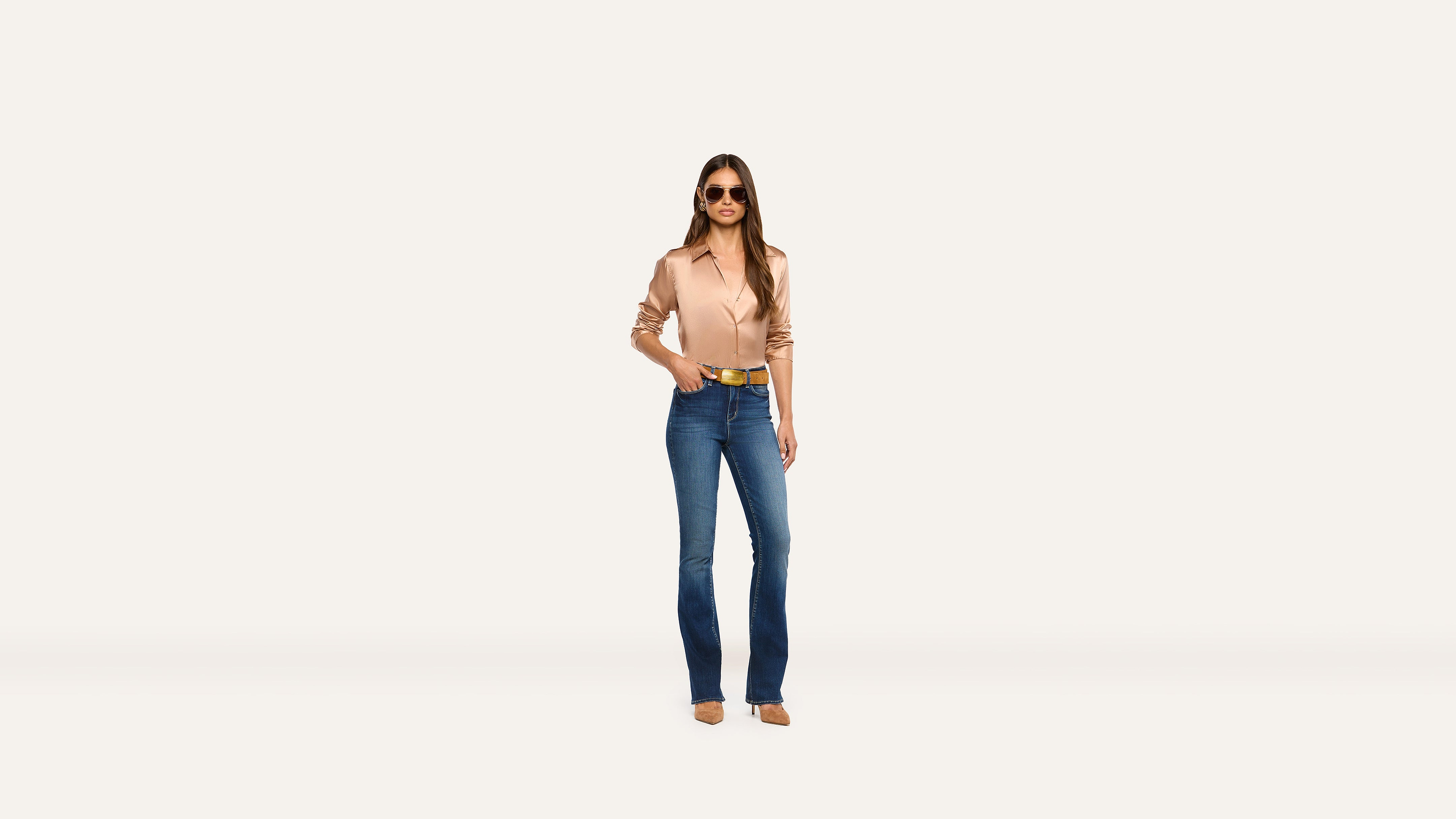 A woman in a tan button-down shirt, wide gold belt, and blue jeans stands against a plain background.