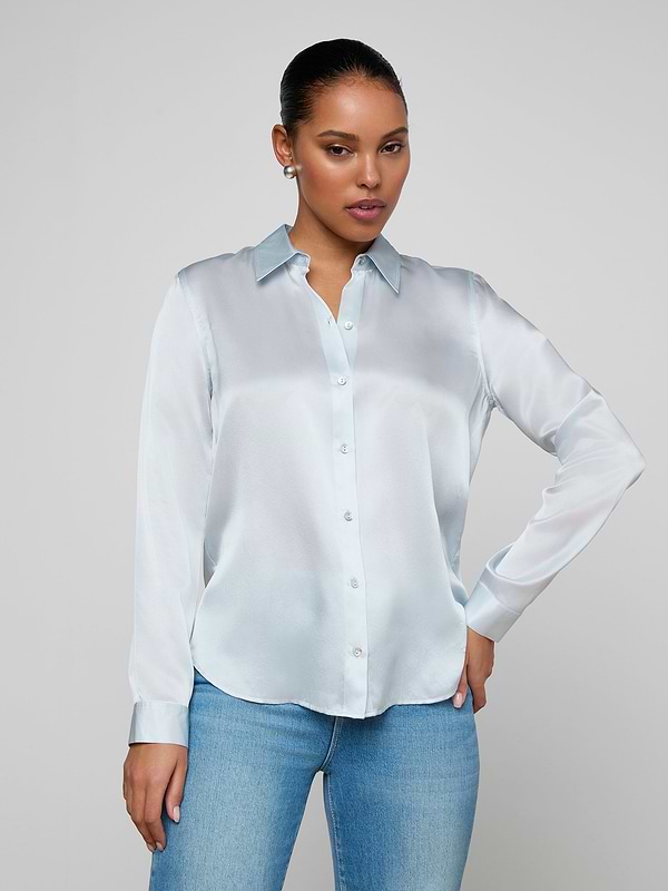 Wearing the Tyler Silk Blouse in pale mist blue and a pair of blue jeans, a person poses against a plain light background with one hand on their hip and the other relaxed by their side.