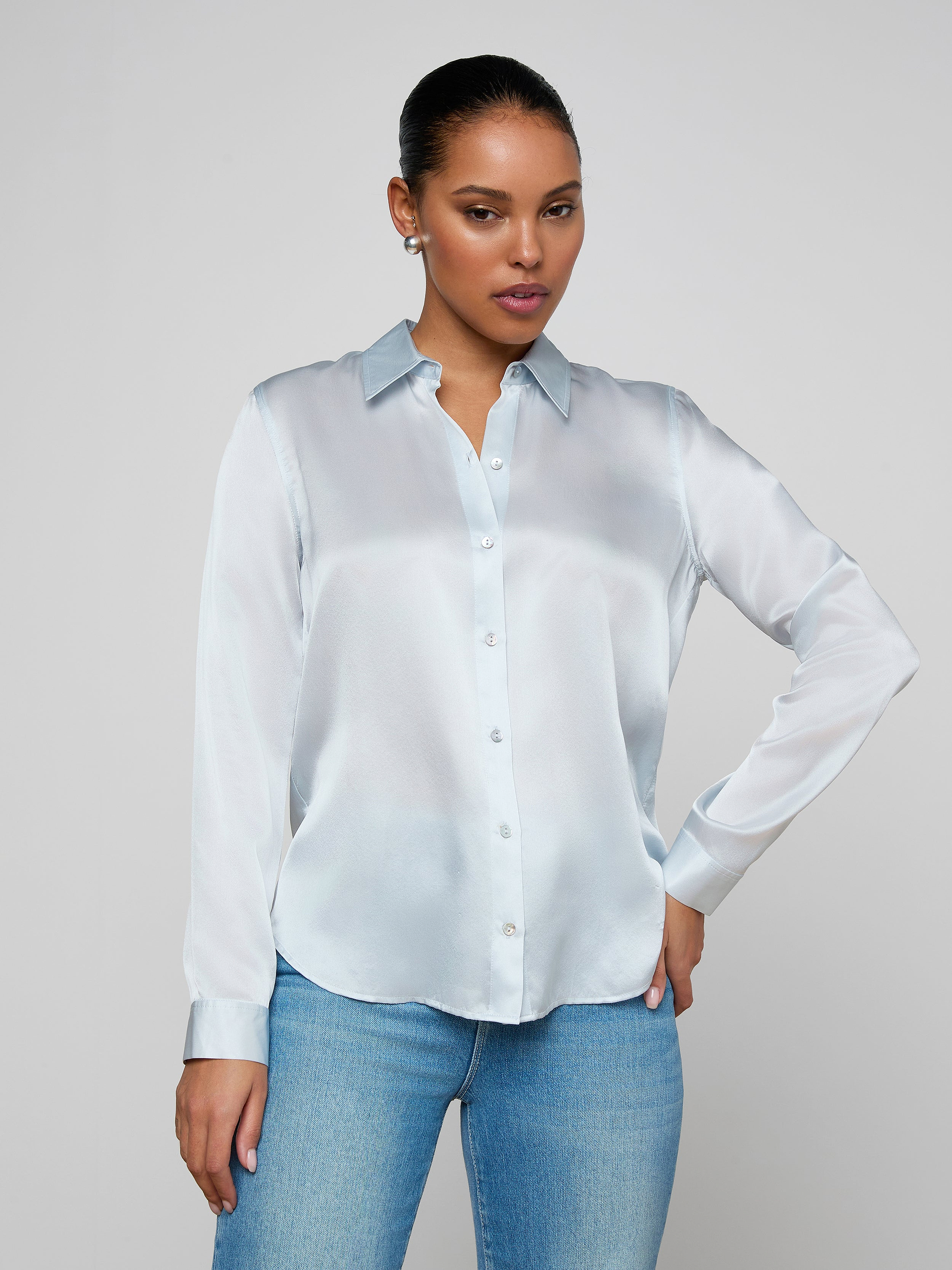 Wearing the Tyler Silk Blouse in pale mist blue and a pair of blue jeans, a person poses against a plain light background with one hand on their hip and the other relaxed by their side.
