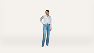 A woman stands against a white background, wearing a white button-down shirt, blue jeans, and heels. She has one hand on her hip and looks to the side.