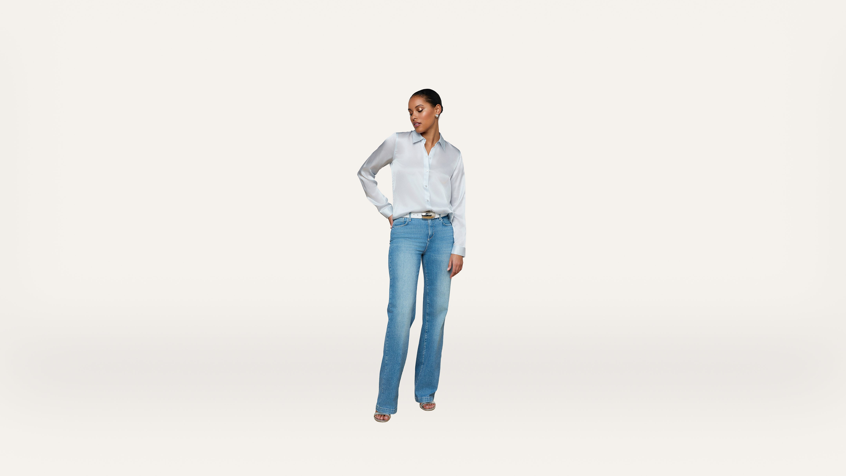 A woman stands against a white background, wearing a white button-down shirt, blue jeans, and heels. She has one hand on her hip and looks to the side.