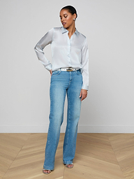 A woman stands against a plain white background, wearing the Tyler Silk Blouse in pale mist blue, tucked into high-waisted light blue jeans with open-toe heels. She has one hand in her pocket and gazes slightly downward to her left.