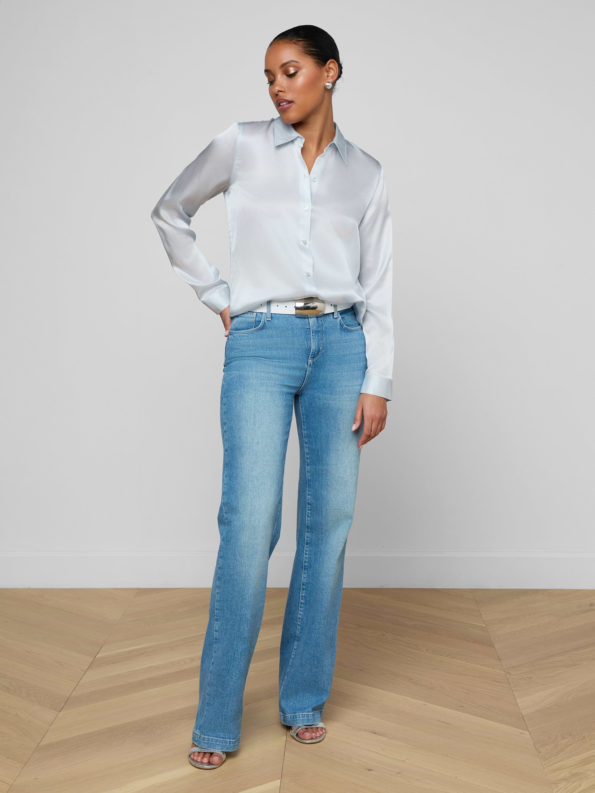 A woman stands against a plain white background, wearing the Tyler Silk Blouse in pale mist blue, tucked into high-waisted light blue jeans with open-toe heels. She has one hand in her pocket and gazes slightly downward to her left.