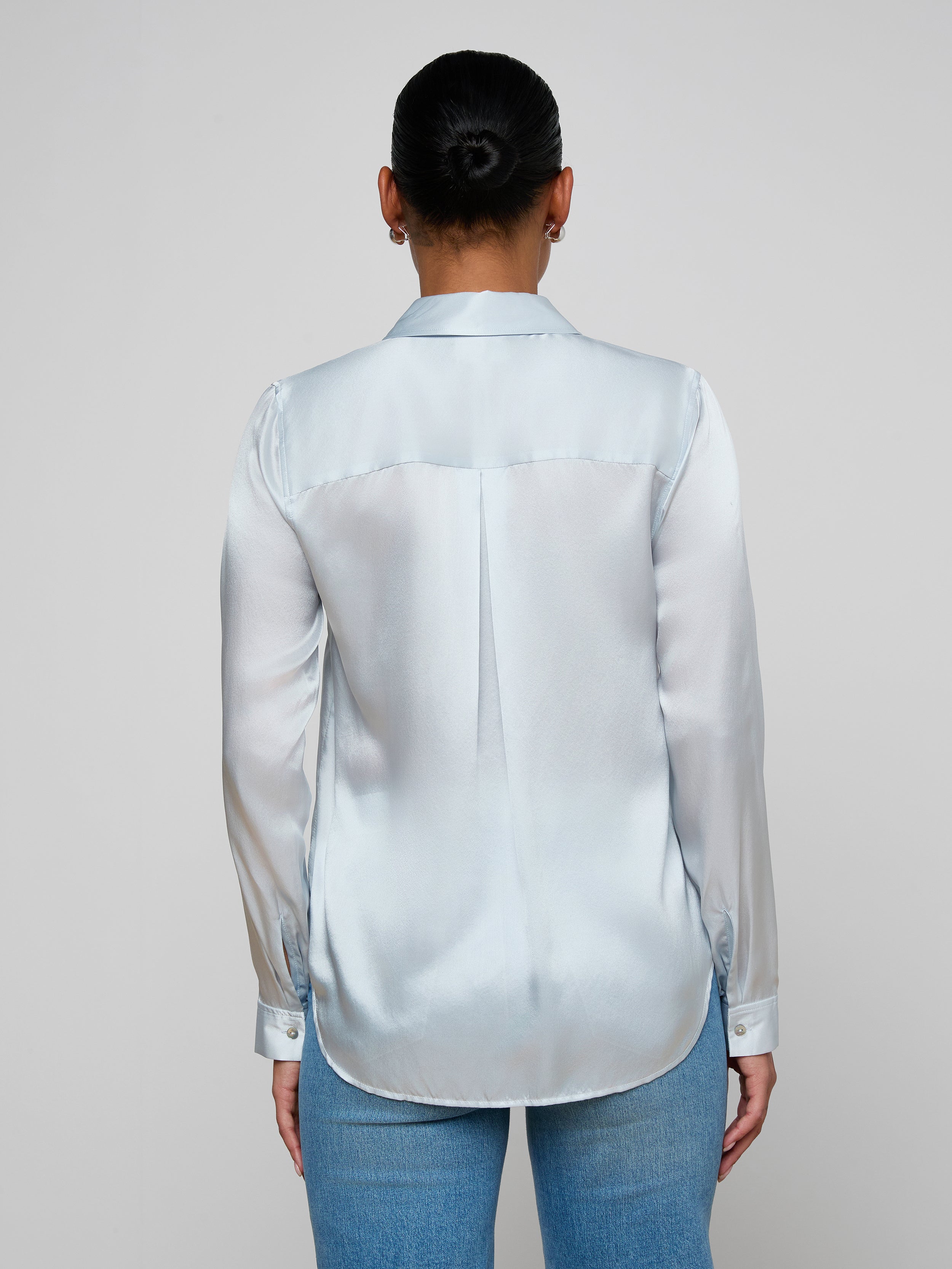 A person with dark hair in a bun wears the Tyler Silk Blouse in pale mist blue and blue jeans, standing with their back to the camera against a plain light background.