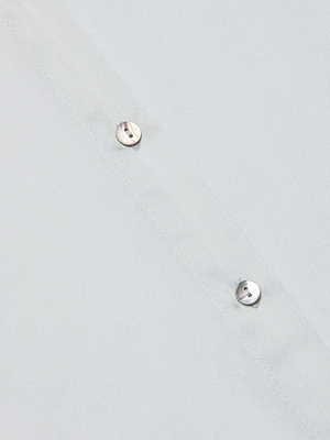 Close-up of the Tyler Silk Blouse in light grey—a smooth, slightly shiny button-down with a seam and two small white buttons sewn diagonally, reflecting L'AGENCE’s signature elegance.