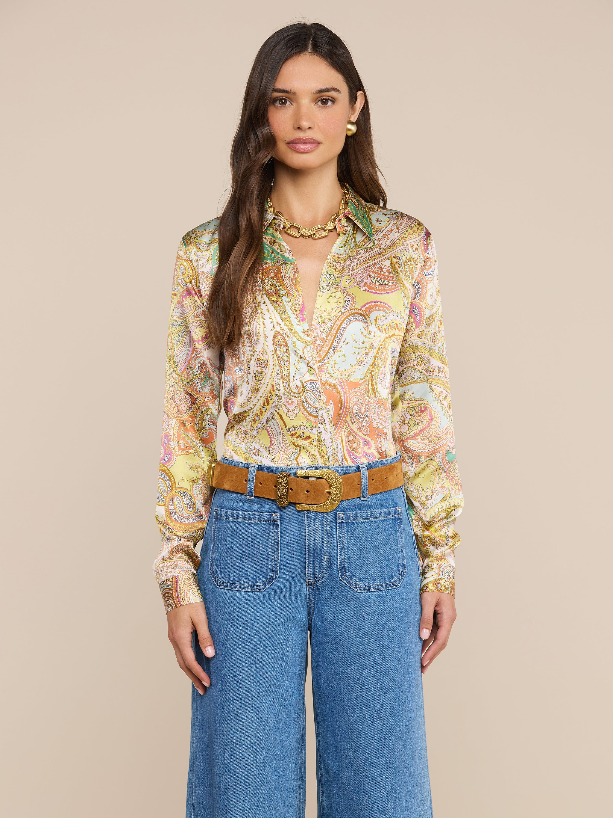Eine Frau mit langen braunen Haaren trägt eine bunte Tyler-Seidenbluse mit Paisley-Muster, eine blaue Jeans mit hoher Taille, einen hellbraunen Wildledergürtel, eine goldene Halskette und goldene Ohrringe und steht vor einem schlichten beigen Hintergrund.