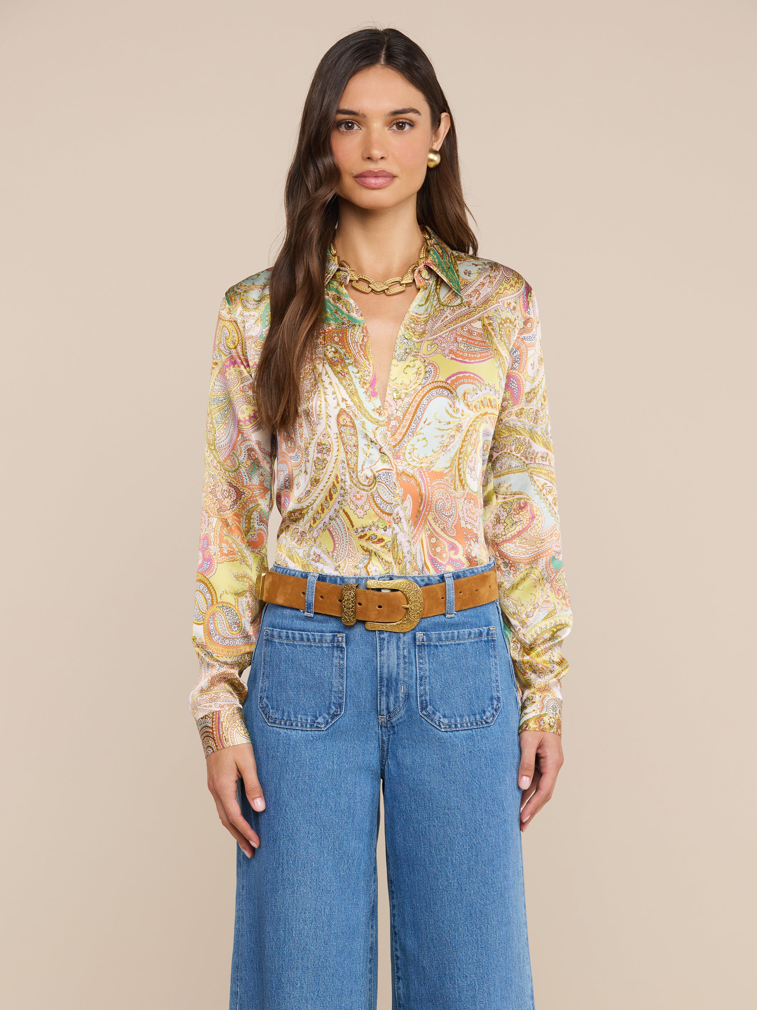Eine Frau mit langen braunen Haaren trägt eine bunte Tyler-Seidenbluse mit Paisley-Muster, eine blaue Jeans mit hoher Taille, einen hellbraunen Wildledergürtel, eine goldene Halskette und goldene Ohrringe und steht vor einem schlichten beigen Hintergrund.