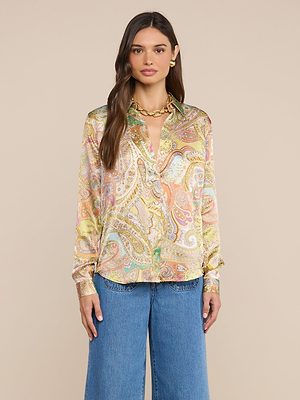 A woman with long brown hair and a neutral expression stands against a beige background, wearing the Tyler Silk Blouse in a colorful paisley print, blue jeans, and a gold chain necklace.