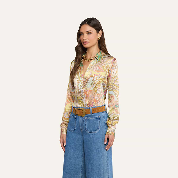 Eine Frau mit langen, welligen braunen Haaren posiert vor einem beigen Hintergrund. Sie trägt die „Tyler Silk Blouse“ von L’AGENCE mit buntem Paisley-Muster, eine blaue Jeans mit weitem Bein, einen hellbraunen Gürtel mit großer Schnalle, goldene Ohrringe und eine klobige goldene Halskette.