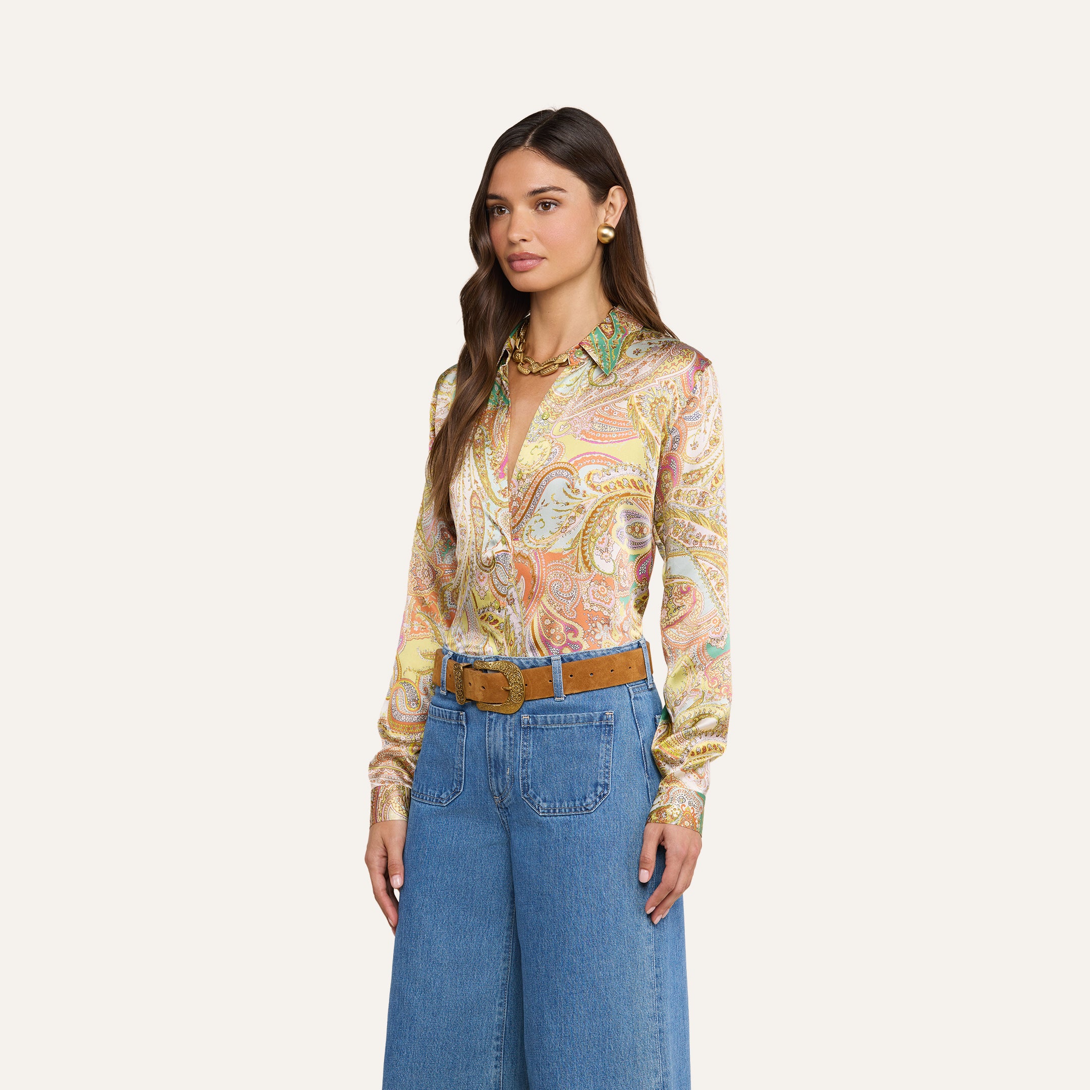 Eine Frau mit langen, welligen braunen Haaren posiert vor einem beigen Hintergrund. Sie trägt die „Tyler Silk Blouse“ von L’AGENCE mit buntem Paisley-Muster, eine blaue Jeans mit weitem Bein, einen hellbraunen Gürtel mit großer Schnalle, goldene Ohrringe und eine klobige goldene Halskette.