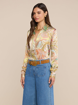 A woman with long wavy brown hair poses against a beige background wearing the L’AGENCE Tyler Silk Blouse in colorful paisley print, wide-leg blue jeans, a tan belt with a large buckle, gold earrings, and a chunky gold necklace.