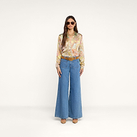 Vor einem schlichten, hellbeigen Hintergrund steht eine Frau mit Sonnenbrille, die die Tyler-Seidenbluse mit Paisley-Muster, eine blaue Jeans mit weitem Bein, einen braunen Gürtel, beige Pumps und eine goldene Halskette trägt.