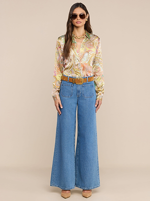 A woman in sunglasses, the Tyler Silk Blouse with a paisley print, wide-leg blue jeans and brown belt, beige heels, and a gold necklace stands against a plain light beige background.