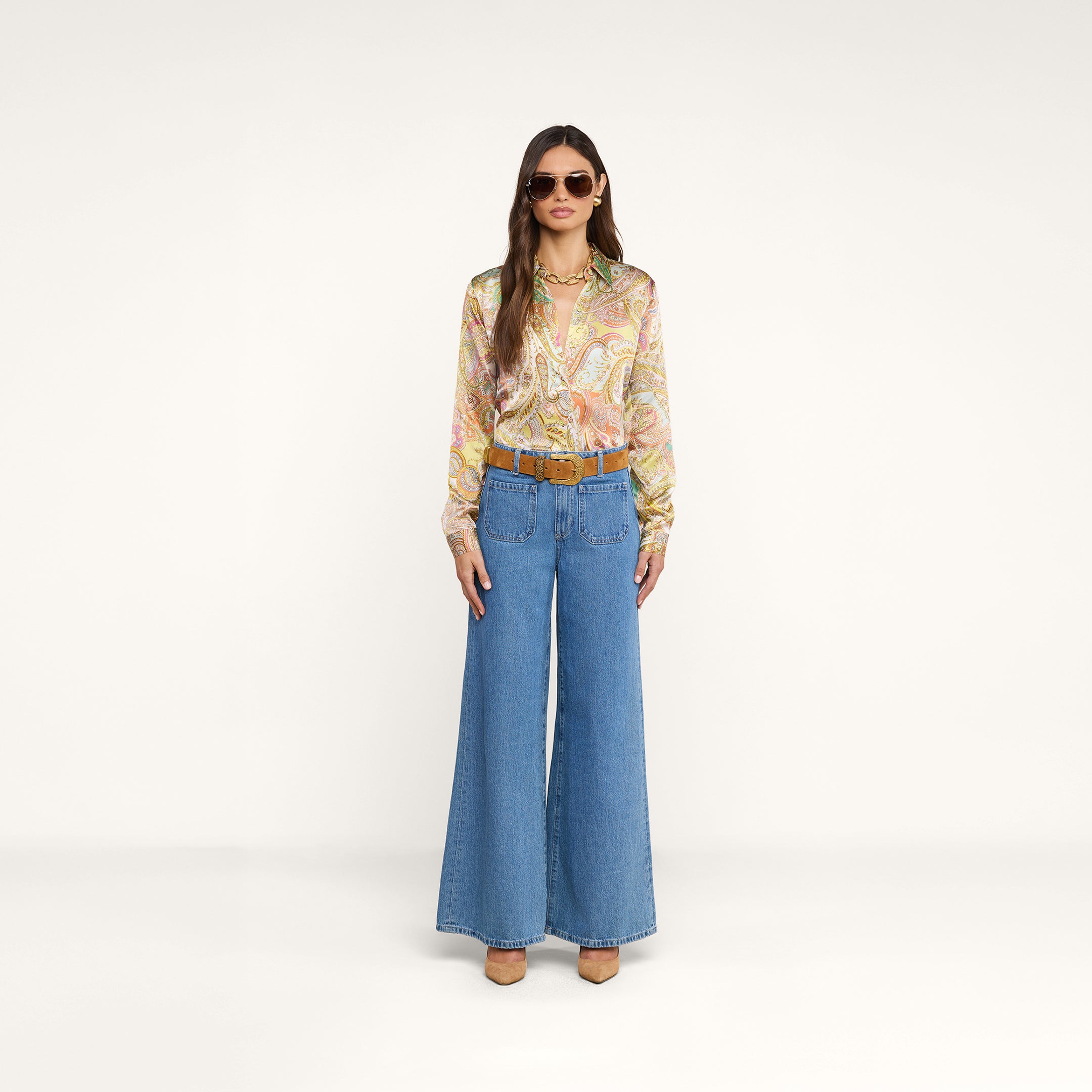 Vor einem schlichten, hellbeigen Hintergrund steht eine Frau mit Sonnenbrille, die die Tyler-Seidenbluse mit Paisley-Muster, eine blaue Jeans mit weitem Bein, einen braunen Gürtel, beige Pumps und eine goldene Halskette trägt.
