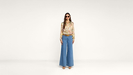 A woman wearing sunglasses, a colorful patterned blouse, blue high-waisted wide-leg jeans, brown shoes, and a belt stands facing forward against a plain white background.