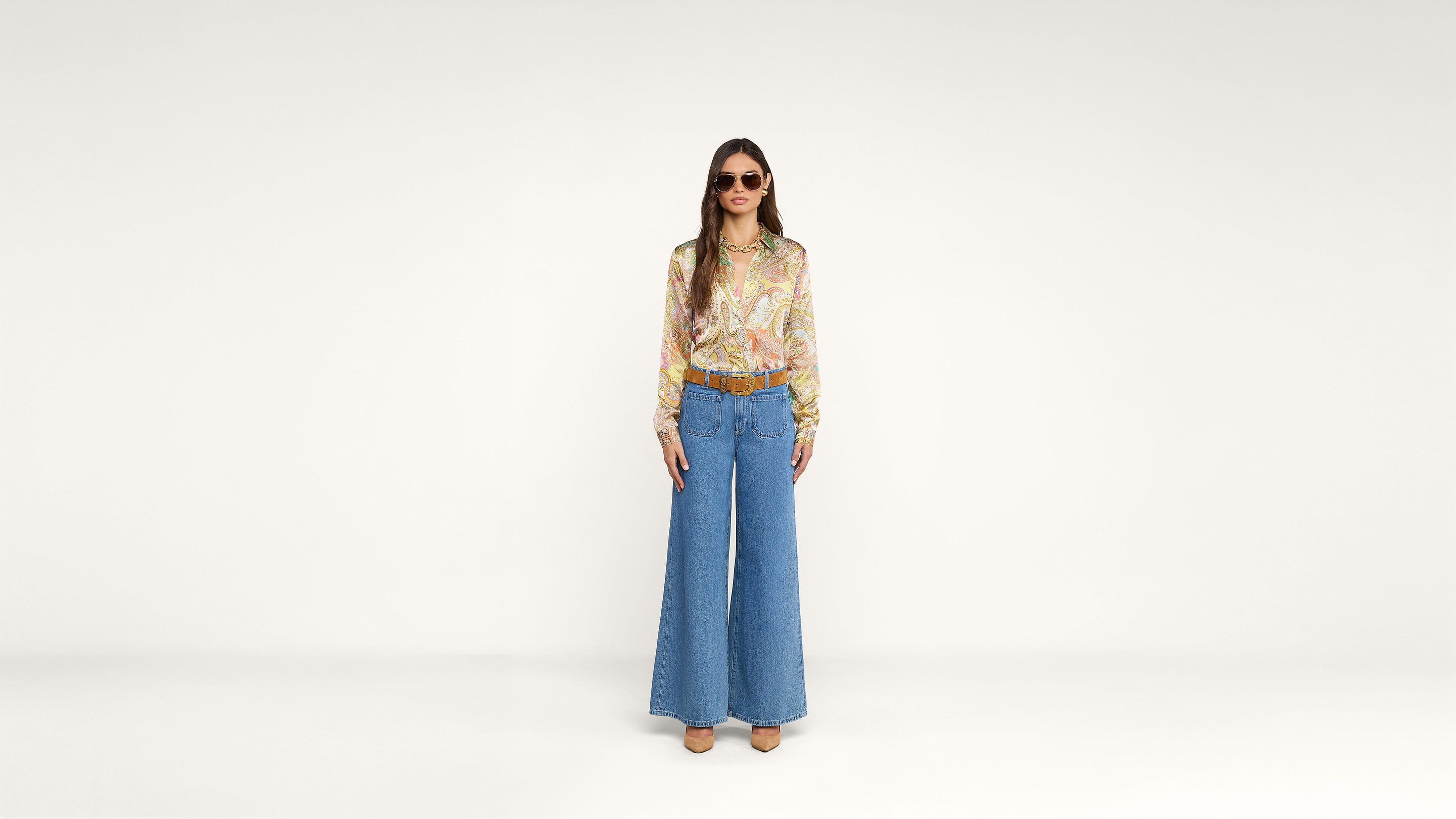 A woman wearing sunglasses, a colorful patterned blouse, blue high-waisted wide-leg jeans, brown shoes, and a belt stands facing forward against a plain white background.