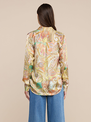 A person with long brown hair, seen from behind, wears the vibrant Tyler Silk Blouse in a paisley print and blue wide-leg jeans against a beige background.