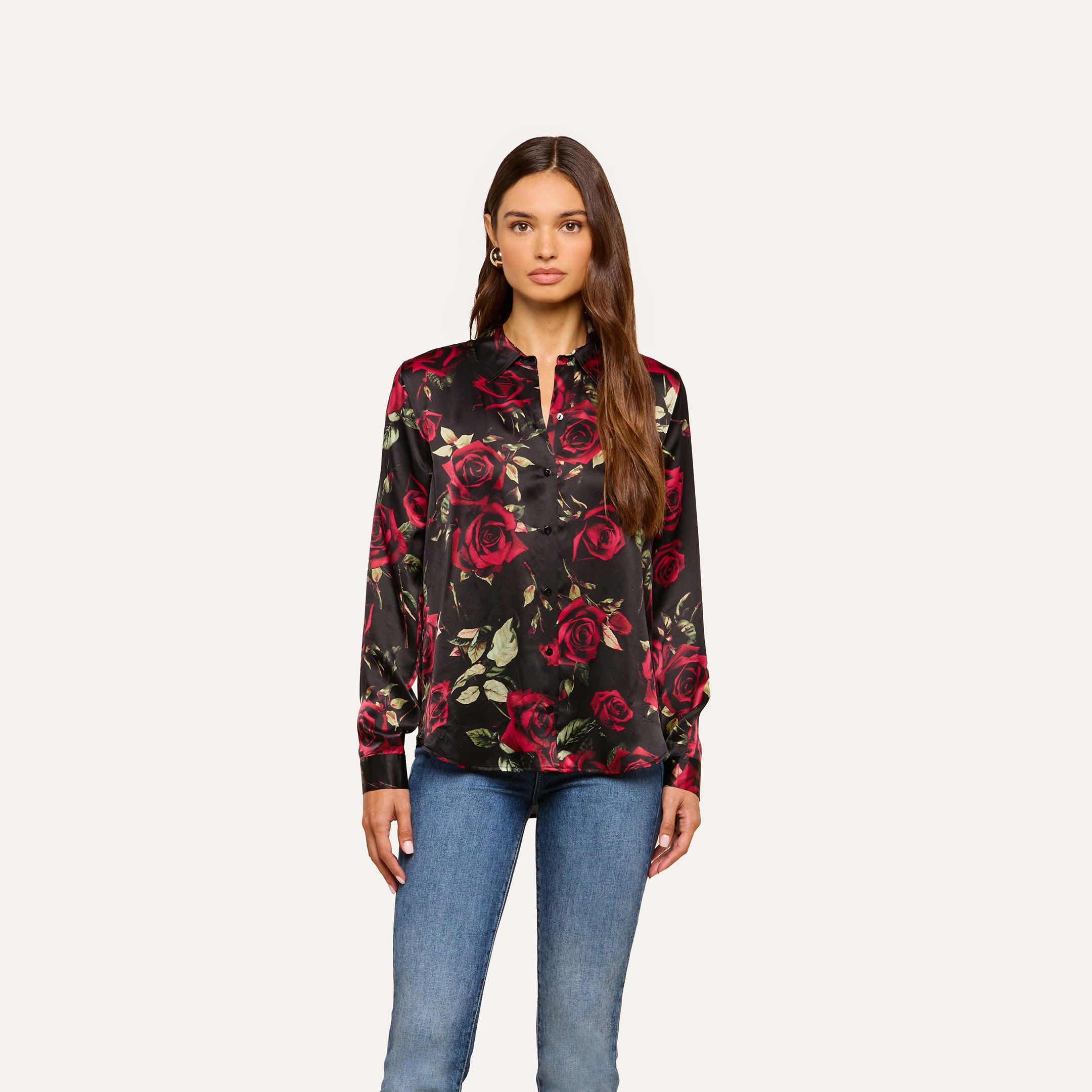 A woman stands against a plain background, wearing the Tyler Silk Blouse—featuring a black silk fabric adorned with a red and green rose print—paired with blue jeans. Her long hair is loose, and she faces forward with a neutral expression, showcasing an elegant option for the business wardrobe.