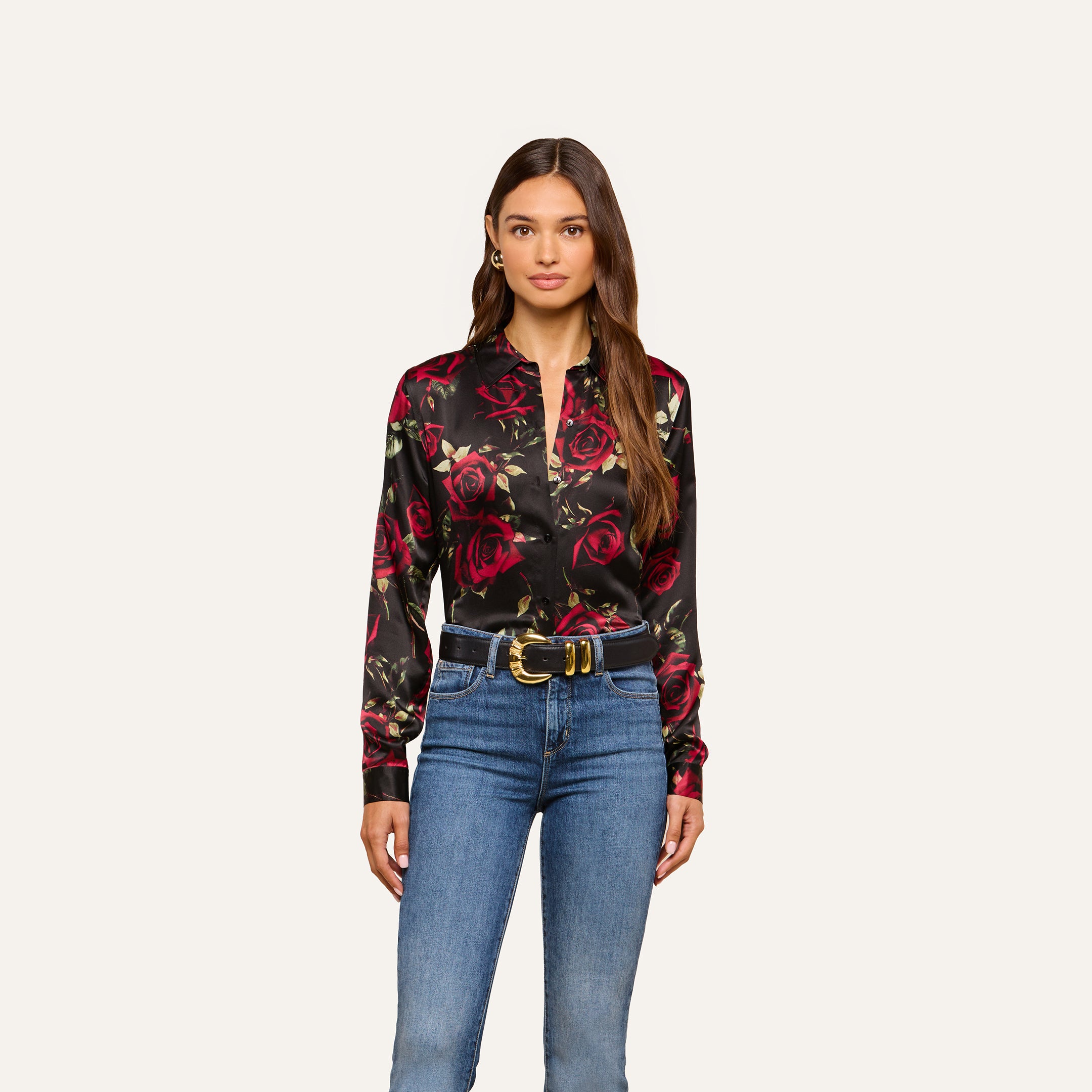 A woman stands facing forward against a plain light background, wearing the Tyler Silk Blouse—a black silk shirt with a red rose print—paired with blue jeans and a black belt with a gold buckle.