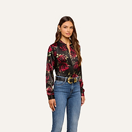 A woman with long brown hair and a neutral expression stands against a plain background, wearing the Tyler Silk Blouse featuring red rose prints, blue jeans, a black belt with a gold buckle, and earrings. This look adds elegance to any business wardrobe.