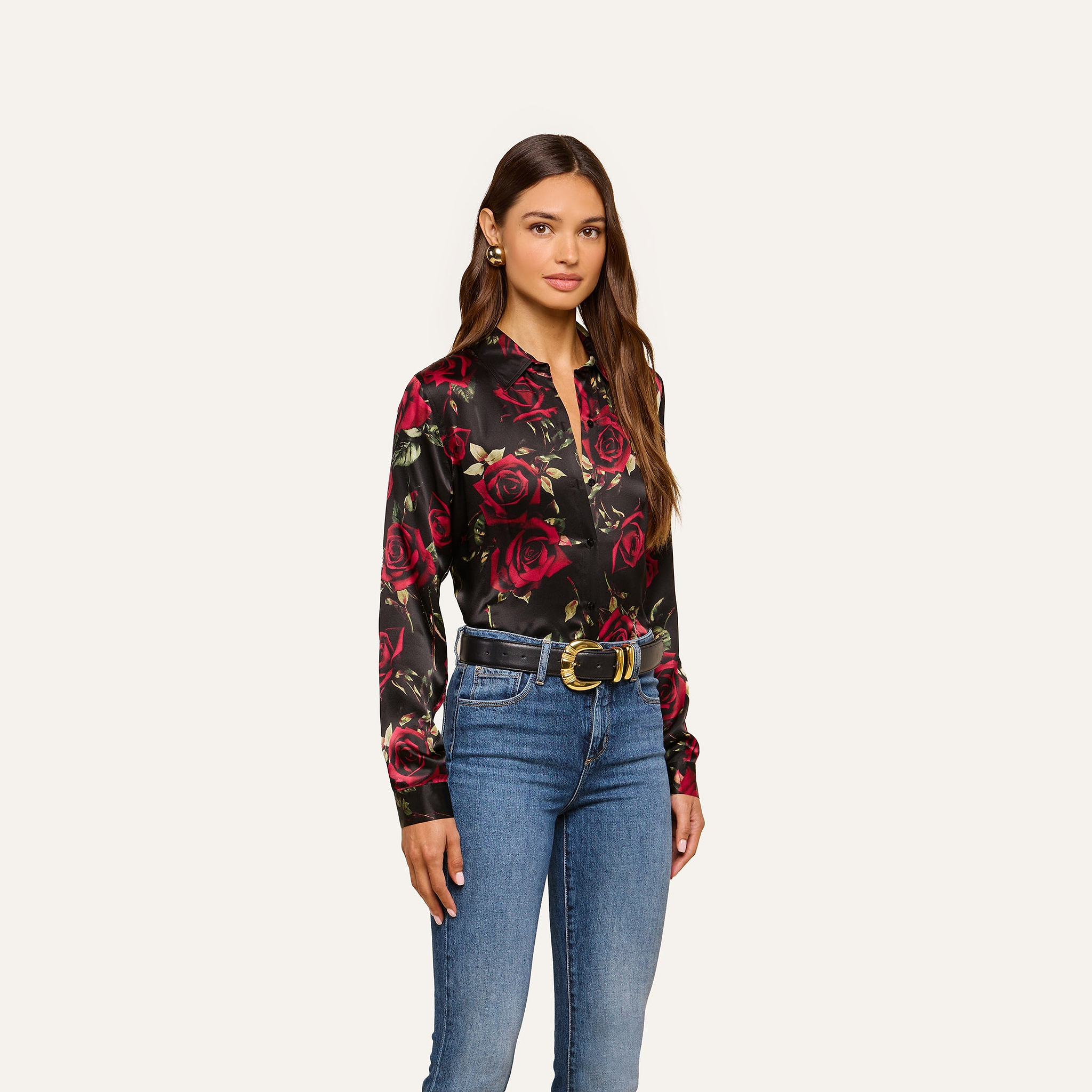 A woman with long brown hair and a neutral expression stands against a plain background, wearing the Tyler Silk Blouse featuring red rose prints, blue jeans, a black belt with a gold buckle, and earrings. This look adds elegance to any business wardrobe.