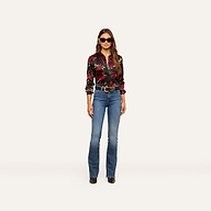 A woman wearing sunglasses, the Tyler Silk Blouse with a black rose print, blue jeans, a belt, and black shoes stands facing forward against a plain, light background.