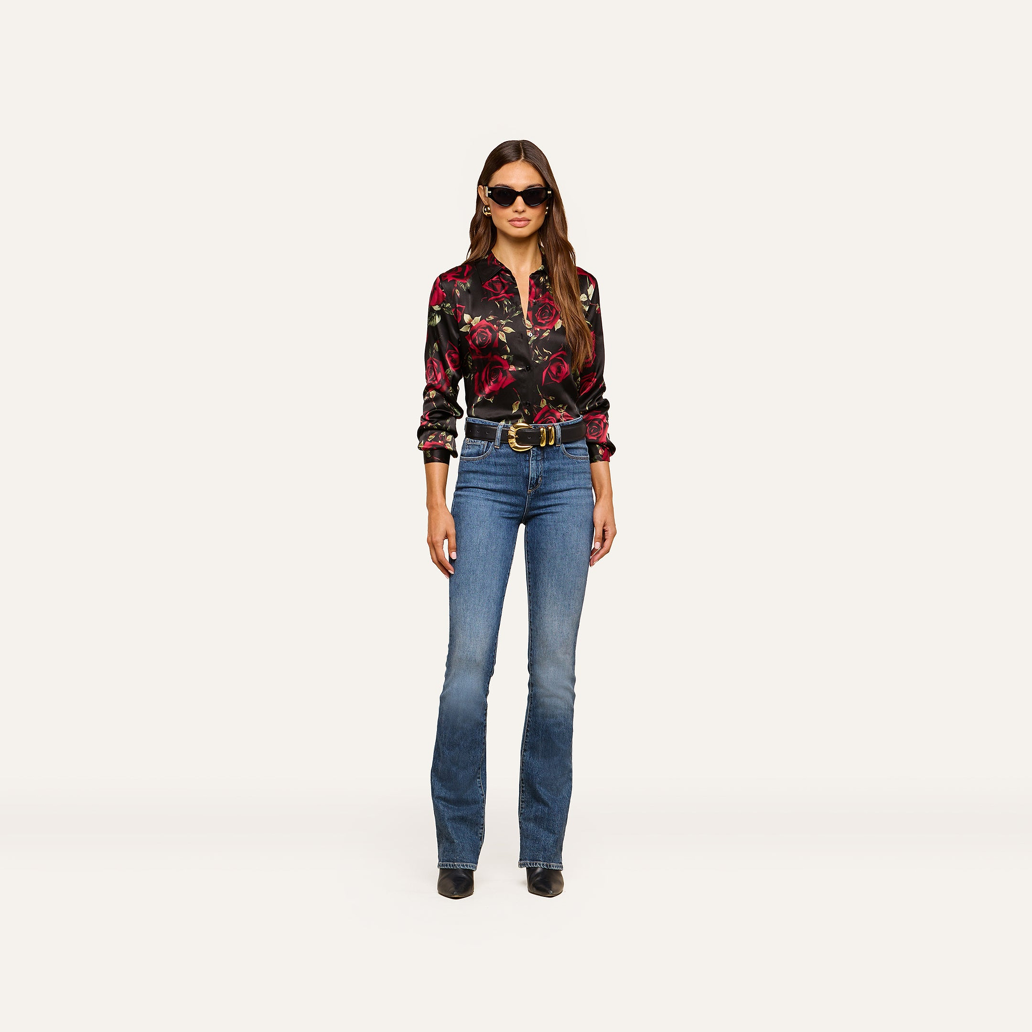 A woman wearing sunglasses, the Tyler Silk Blouse with a black rose print, blue jeans, a belt, and black shoes stands facing forward against a plain, light background.