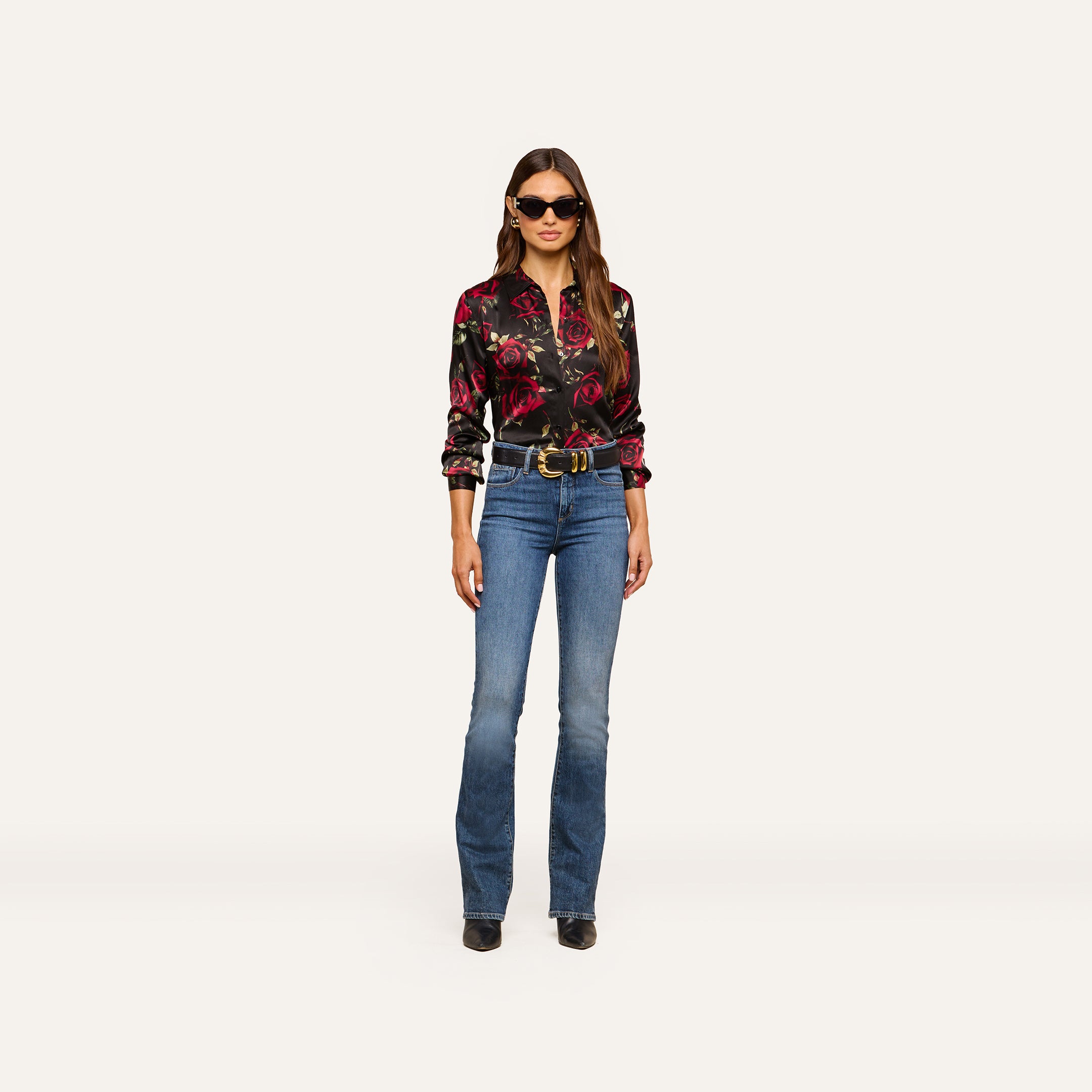 A woman wearing sunglasses, the Tyler Silk Blouse with a black rose print, blue jeans, a belt, and black shoes stands facing forward against a plain, light background.