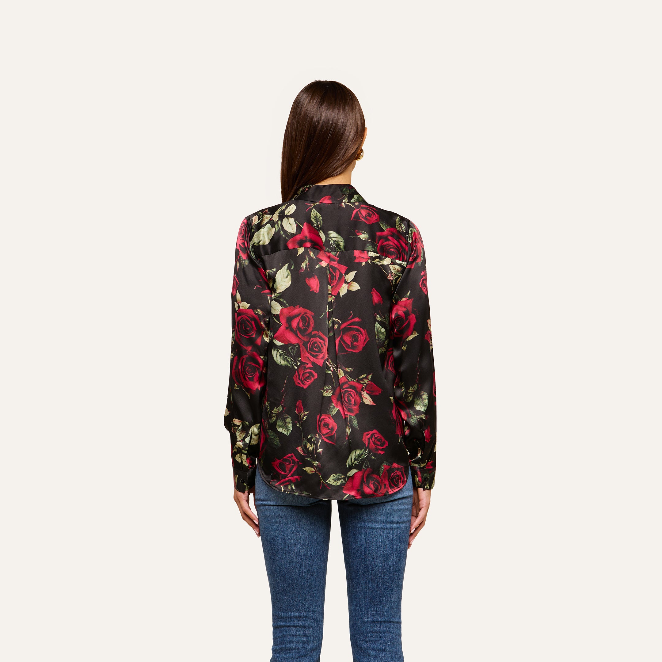 A woman with long brown hair wears the Tyler Silk Blouse, featuring a red rose print, paired with blue jeans. She stands with her back to the camera against a plain light background.