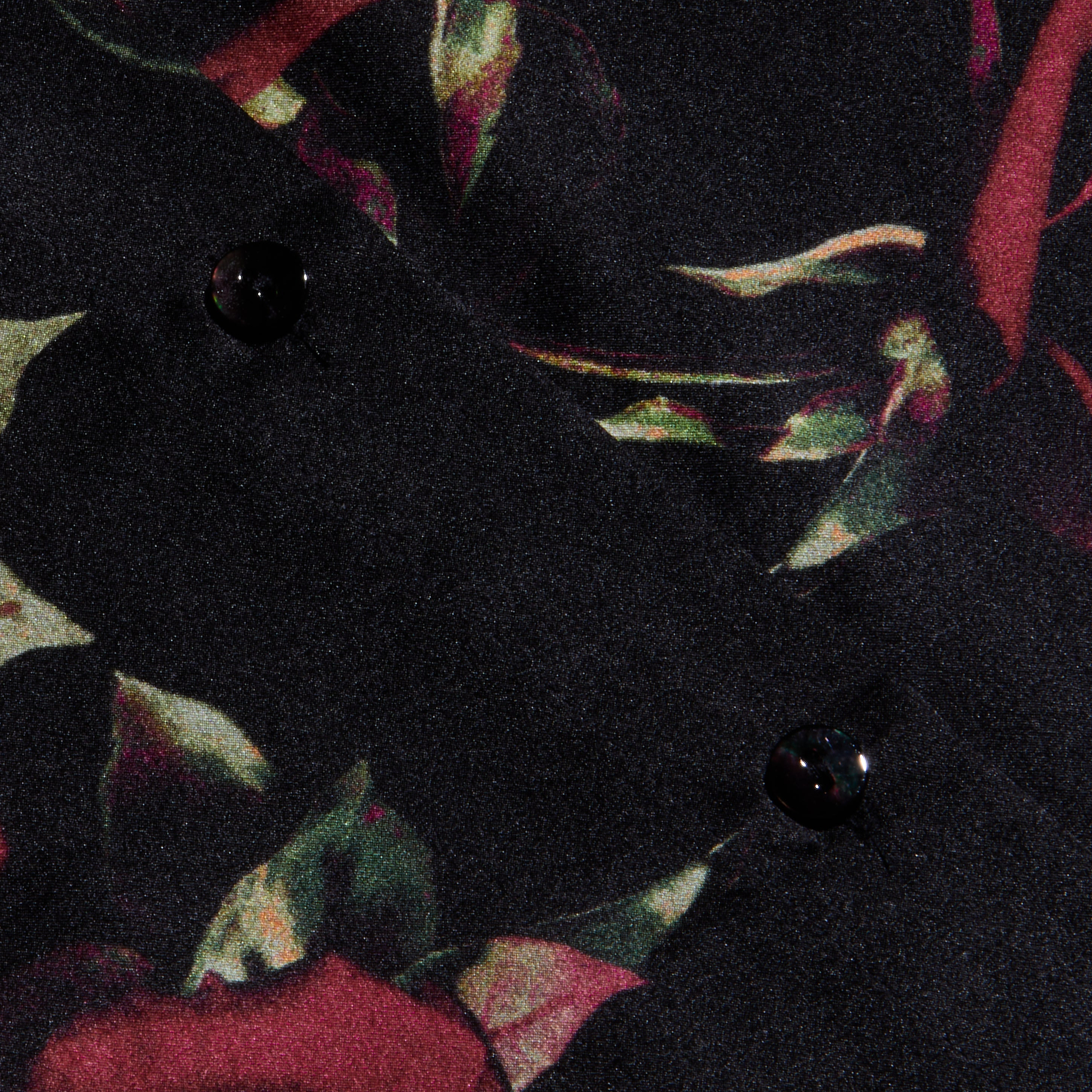 Close-up of the Tyler Silk Blouse fabric, showcasing a dark floral pattern with red and green leaves reminiscent of a rose print, accented by two black buttons. The material appears soft and slightly textured, making it a refined addition to your business wardrobe.
