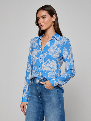 A woman with long brown hair wears the Holly Blouse in regatta blue and white paisley print, styled with blue jeans, standing with one hand in her pocket against a plain light gray background.