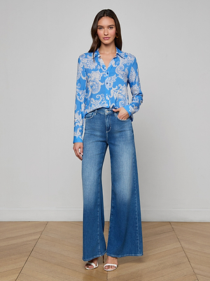 A woman with long brown hair poses indoors against a plain light background, wearing the Holly Blouse in regatta blue paisley print with long sleeves and button-down front, paired with high-waisted wide-leg blue jeans and silver open-toe heels.