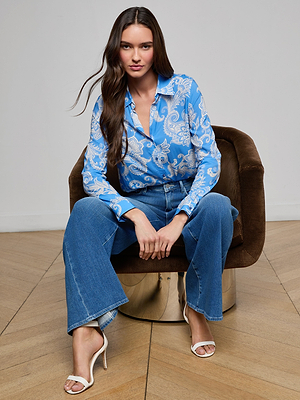 A woman with long brown hair sits on a brown chair, wearing a blue and white paisley blouse, blue flared jeans, and white heeled sandals, with her hands resting on her knees.