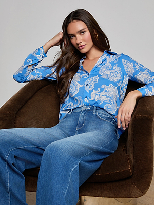 A woman with long brown hair sits in a brown armchair, wearing the Holly Blouse—a regatta blue paisley print long sleeve button down—paired with wide-leg blue jeans. She relaxes, resting her head on her hand and gazing at the camera.
