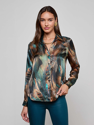 A woman with long brown hair wears gold jewelry, teal pants, and the L'AGENCE Tyler Silk Blouse featuring a teal, brown, and black peacock feather print, standing against a plain light gray background.
