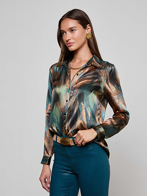A woman with long brown hair wears the Tyler Silk Blouse by L'AGENCE, featuring a shiny multicolored feather print. She pairs it with teal pants, a gold belt, gold earrings, and a gold necklace, standing against a light gray background.