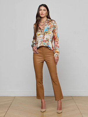 A woman stands indoors against a plain wall, wearing a colorful blouse, the Ketra Leather Bootcut Trouser in tan lambskin leather, beige high-heeled shoes, and layered necklaces. She has long straight brown hair and a neutral expression.