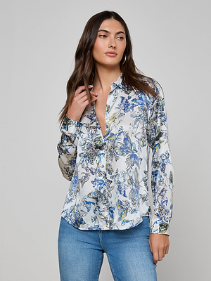 A woman with long brown hair wears the Tyler Silk Blouse with a blue butterfly print and blue jeans, standing against a plain gray background and gazing slightly to the side.