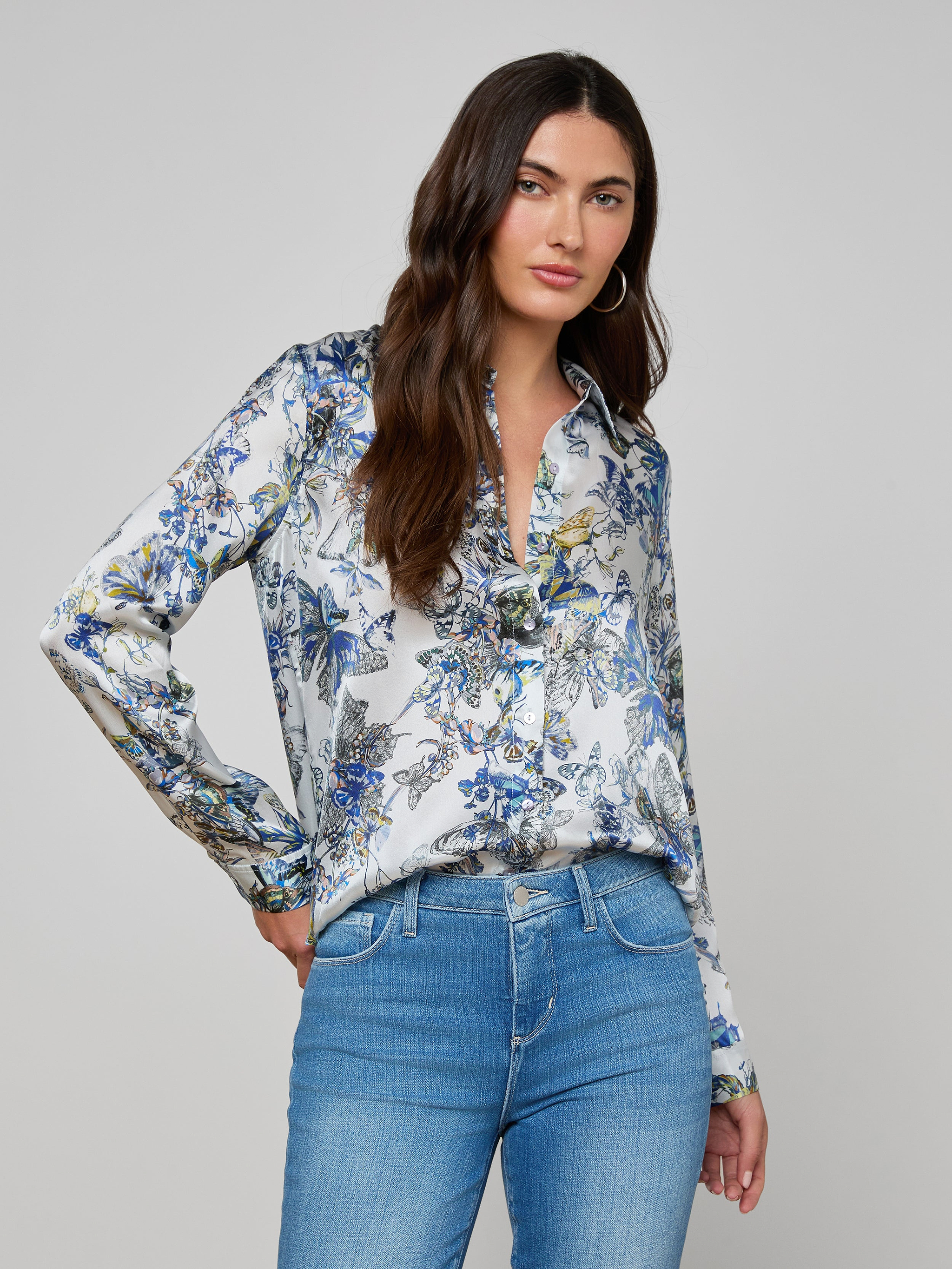 Woman with long brown hair wearing the LAGENCE Tyler Silk Blouse and blue jeans, standing with one hand in her pocket against a plain light gray background.