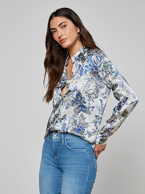 A woman with long brown hair, wearing the Tyler Silk Blouse in a blue and white butterfly print and light blue jeans, stands with one hand in her pocket against a plain grey background.