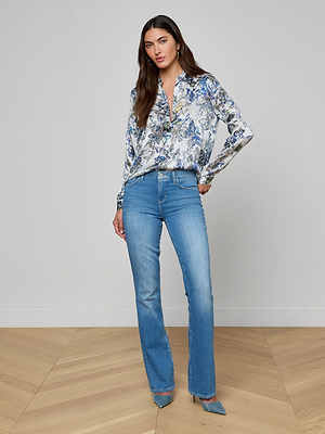 A woman stands indoors on a wooden floor, wearing the Tyler Silk Blouse with a blue and gray butterfly print, light blue jeans, and blue pointed-toe heels. She poses with one hand on her hip in front of a plain white wall.