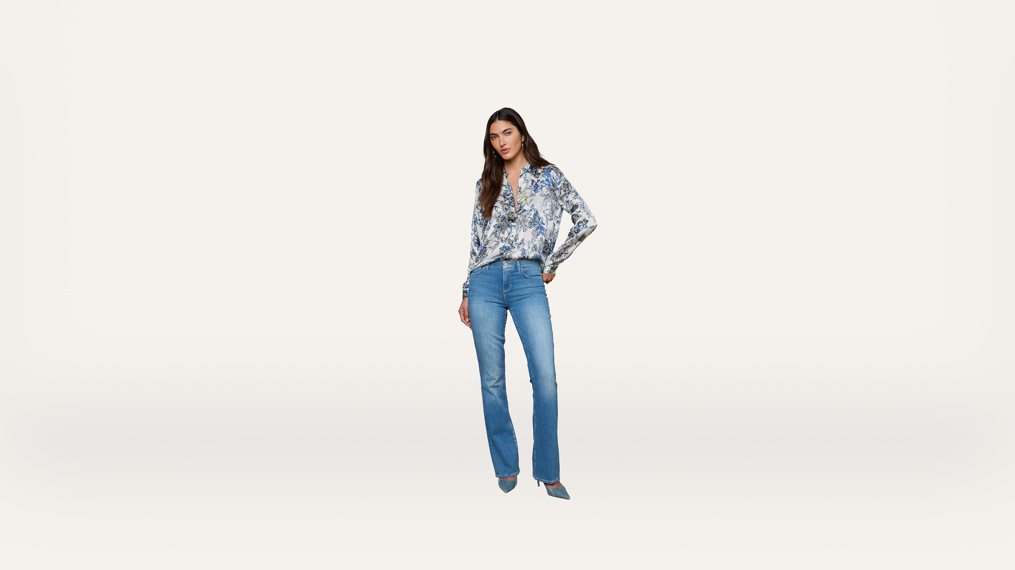 Woman standing on a white background, wearing a floral blouse, blue jeans, and blue heels, with one hand on her hip and a neutral expression.