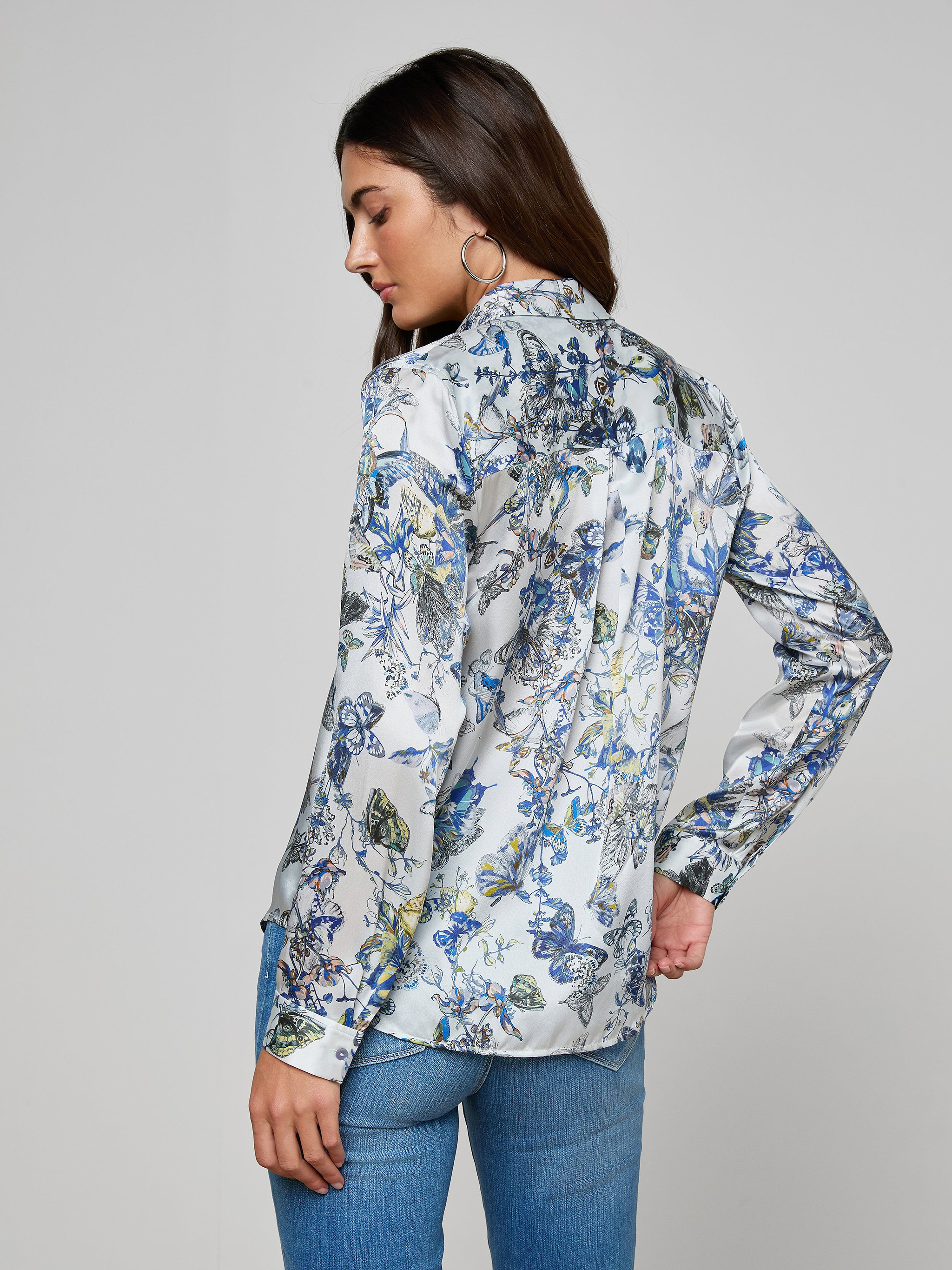 A woman with long brown hair and hoop earrings wears the white Tyler Silk Blouse with a blue and yellow butterfly print and blue jeans, standing against a plain gray background, looking down over her shoulder.
