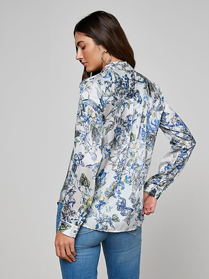 A woman with long brown hair and hoop earrings wears the white Tyler Silk Blouse with a blue and yellow butterfly print and blue jeans, standing against a plain gray background, looking down over her shoulder.